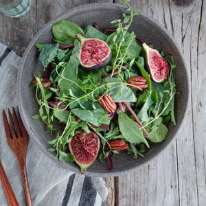 May include: A gray bowl filled with a salad of fresh greens, sliced figs, and pecan halves. The salad is on a rustic wooden table with a glass of water and a bowl of herbs in the background.
