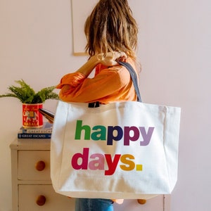 May include: A white canvas tote bag with black handles and the text "happy days." printed in rainbow colours.