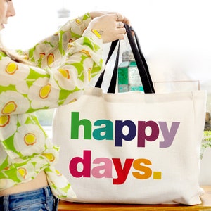 May include: A white canvas tote bag with black handles. The bag has the words "happy days" printed on it in rainbow colours.