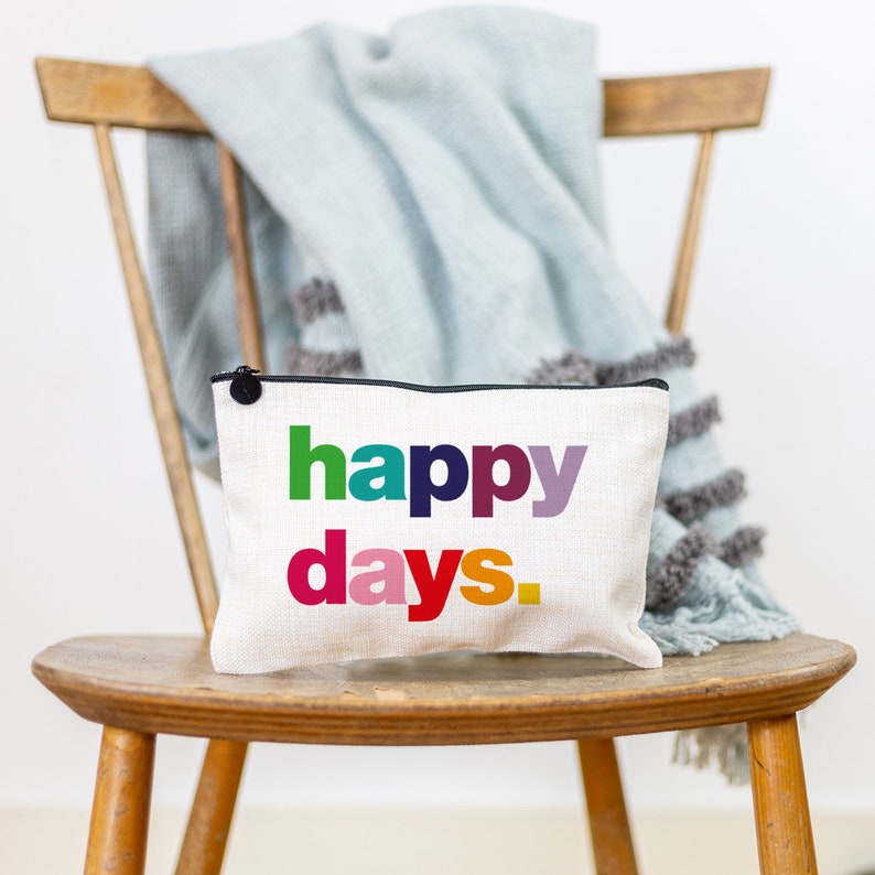 May include: A white canvas zippered pouch with a black zipper and a black button. The pouch has the words "happy days." printed in rainbow colours.