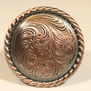 May include: A round, copper-colored metal button with a rope-like border and an intricate, engraved floral design.
