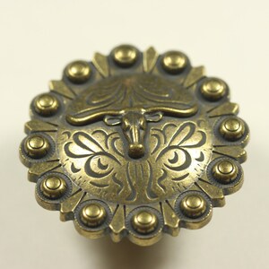 May include: A gold-toned metal belt buckle with a detailed design of a bull's head and floral accents. The buckle is surrounded by a ring of smaller, round metal studs.