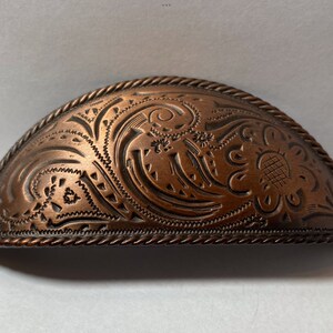 May include: A half-round, copper-colored cabinet pull with a decorative, embossed floral design. The pull has a rope-like edge.