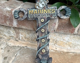 May include: A wooden cross with a rope design and a metal eagle and anchor emblem. The cross has the words "MARINES THE FEW THE PROUD" on it.