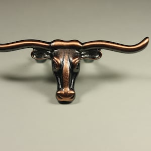 May include: A bronze-colored metal bull head with black horns and a black face. The bull head is a decorative piece with a rustic look.