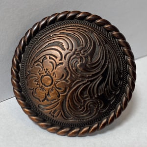 May include: A round, copper-colored metal buckle with a rope-like border and an intricate floral design.