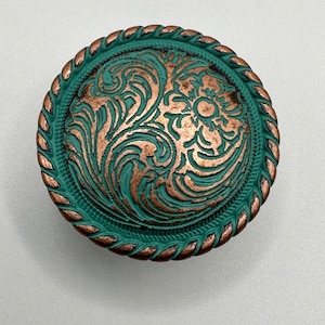 May include: A round, copper-colored metal button with a green, floral design. The button has a rope-like border.