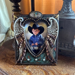 May include: A heart-shaped picture frame with a brown, gold, and turquoise design. The frame features a cross on the top and angel wings on the sides. A photo of a young boy wearing a cowboy hat is inside the frame.