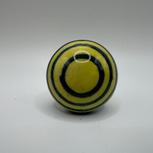 May include: A spherical decorative object with a yellow base and concentric dark blue rings. The object is smooth and appears to be made of ceramic or a similar material. The design is simple and eye-catching.