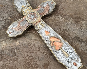 May include: A silver and gold cross with intricate carvings and a star in the center. The cross has a pair of wings on one side and two hearts with an arrow through them on the other side.