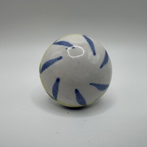 May include: A white ceramic ball with blue and yellow painted stripes. The ball is smooth and has a glossy finish.