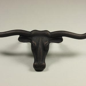 May include: A bronze-colored longhorn steer head sculpture with large, curved horns. The sculpture has a detailed texture and is mounted on a light background. The steer's head is centered, showcasing its features.