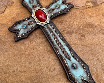 May include: A turquoise and brown leather cross with a red gemstone in the center. The cross is decorated with gold studs and has a distressed finish.