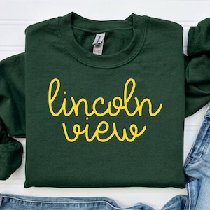 May include: A dark forest green long-sleeve crewneck sweatshirt featuring the yellow cursive text Lincoln View printed across the center of the chest. The garment is paired with light blue distressed denim jeans with a visible silver button.