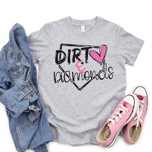 May include: Gray t-shirt with a black baseball diamond graphic and the text "Dirt & Diamonds" with a pink heart. The shirt is paired with a denim jacket and pink sneakers.