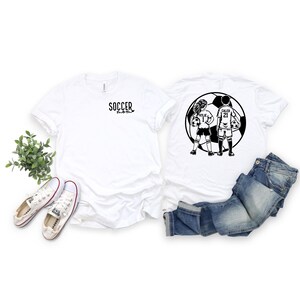 May include: Two white t-shirts with black soccer-themed graphics. One shirt says "Soccer Mom" and the other has a graphic of two children playing soccer with the names "Kylie" and "Caleb" and their jersey numbers.