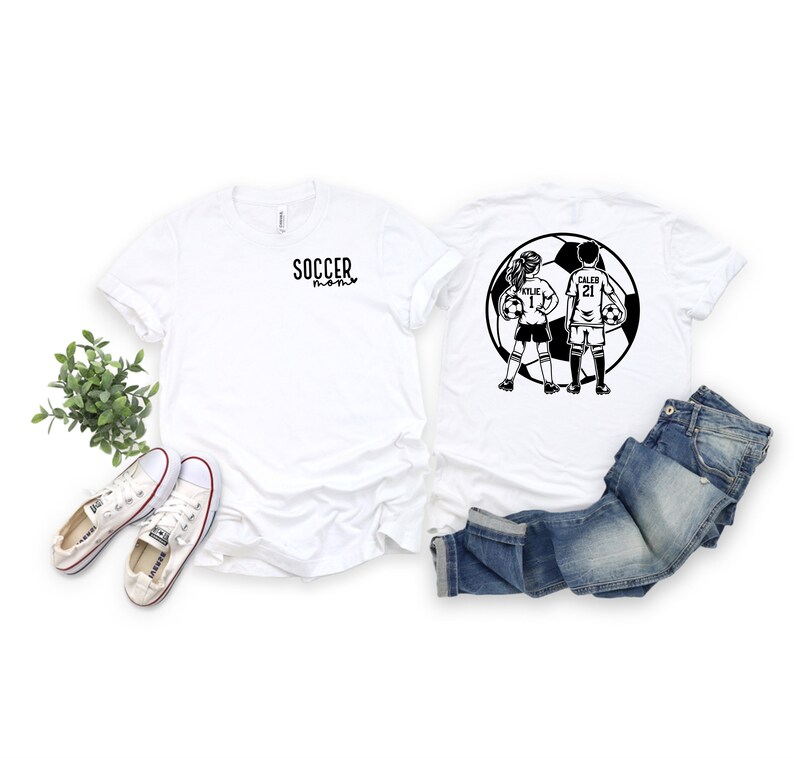 May include: Two white t-shirts with black soccer-themed graphics. One shirt says "Soccer Mom" and the other has a graphic of two children playing soccer with the names "Kylie" and "Caleb" and their jersey numbers.