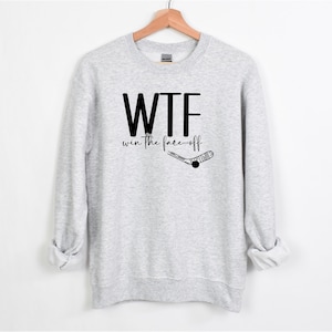May include: A light gray sweatshirt with the text "WTF win the face off" in black. A hockey stick and puck graphic is below the text. The sweatshirt is hanging on a wooden hanger against a white background.