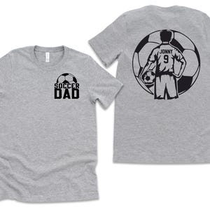 Puede incluir: Dos camisetas grises. Una camiseta tiene la inscripci&oacute;n "SOCCER DAD" en negro con un gr&aacute;fico de bal&oacute;n de f&uacute;tbol. La otra camiseta tiene un gr&aacute;fico de jugador de f&uacute;tbol en la espalda con el nombre "Jonny" y el n&uacute;mero "9".