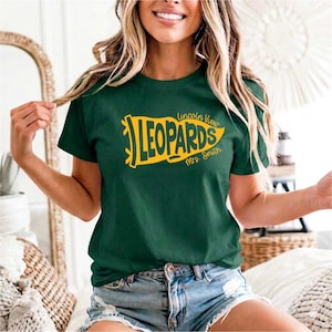 May include: A dark green short-sleeved t-shirt featuring a yellow pennant-shaped graphic on the chest. The graphic contains the text Lincoln View LEOPARDS and Mrs. Smith in a bold, stylized font. The shirt has a crew neckline and a standard fit.