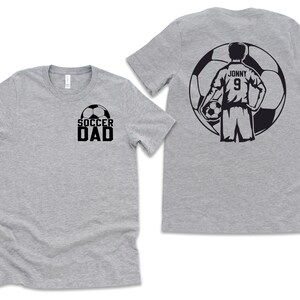 Puede incluir: Dos camisetas grises. Una camiseta tiene la inscripci&oacute;n "SOCCER DAD" en negro con un gr&aacute;fico de bal&oacute;n de f&uacute;tbol. La otra camiseta tiene un gr&aacute;fico de jugador de f&uacute;tbol en la espalda con el nombre "Jonny" y el n&uacute;mero "9".