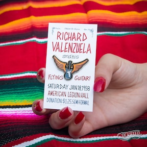 May include: A gold-colored enamel pin featuring a guitar with wings. The pin is on a white card with the text "Richard Valenzuela and his Flying Guitar! Saturday Jan 18, 1958 American Legion Hall Donation $1.25, Semi-Formal DEVOTED3.COM DEVOTED"
