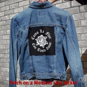 May include: A blue denim jacket with a black patch on the back. The patch features a white rose with the text "Como La Flor Con Tanto Amor" in a circle around it.