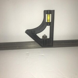May include: A black metal combination square with a level and a ruler. The ruler is marked in inches.
