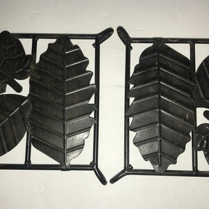 May include: Two black metal leaf-shaped trivets. Each trivet has a square frame with five leaf shapes inside. The leaves are all different sizes and shapes.
