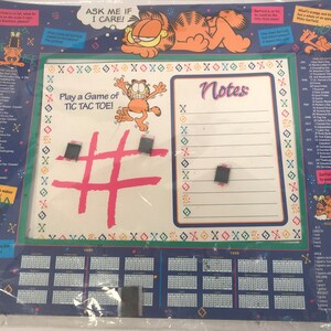 May include: A blue and green Garfield calendar with a white dry-erase board featuring a tic-tac-toe game and a notes section. The board has a pink tic-tac-toe grid and a black hashtag symbol. The calendar features a Garfield cartoon and the text "Ask me if I care!"