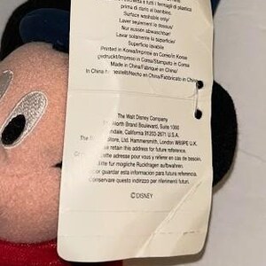 May include: A plush Mickey Mouse toy with a red velvet jacket, black ears, and a tag attached. The tag has text in multiple languages, including English, and the Disney logo.