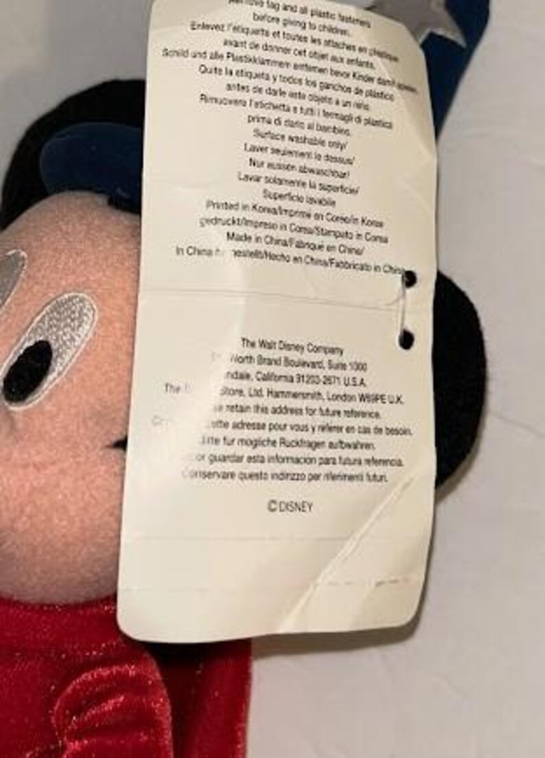 May include: A plush Mickey Mouse toy with a red velvet jacket, black ears, and a tag attached. The tag has text in multiple languages, including English, and the Disney logo.