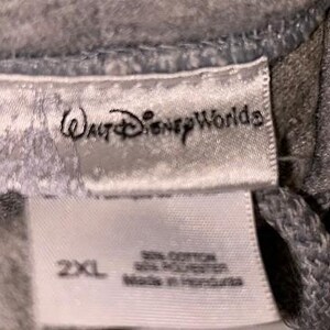 May include: Close-up of a gray garment tag with the text "Walt Disney World" in a script font. Below the logo, the tag reads "2XL" and lists the fabric content and country of origin.