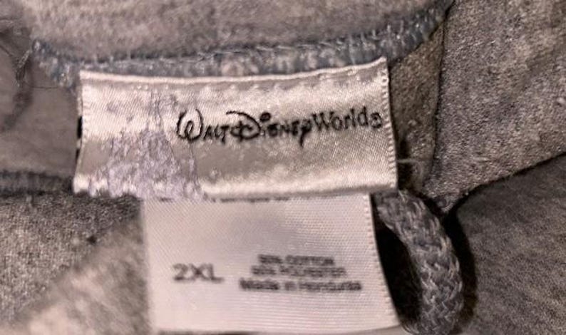 May include: Close-up of a gray garment tag with the text "Walt Disney World" in a script font. Below the logo, the tag reads "2XL" and lists the fabric content and country of origin.