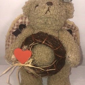 May include: A brown teddy bear wearing a plaid fabric angel wing costume and holding a brown twig wreath with a red heart attached.