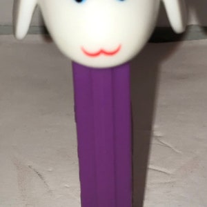 May include: A purple plastic dispenser with a white lamb head on top. The lamb has a red bow, blue eyes, and a pink mouth.