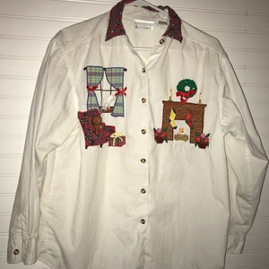 May include: A white long-sleeved button-down shirt with a red and green patterned collar. The shirt features embroidered Christmas scenes, including a fireplace, a window, and a teddy bear sitting in a chair.