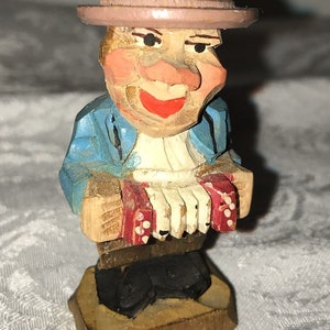 May include: A wooden figurine of a man wearing a brown hat and blue jacket, playing an accordion. The figurine is painted with details and has a smiling face.
