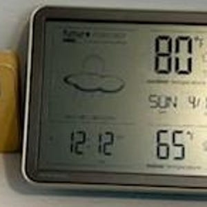 May include: A digital weather station with a silver frame displays the temperature as 80°F, the date, and time. A smaller, yellow, rectangular sensor is to the left. The screen also shows the current humidity.