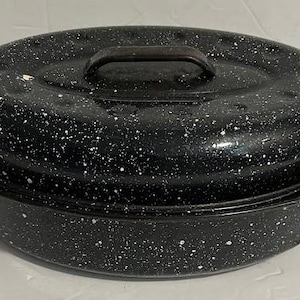 May include: A black oval roasting pan with a lid. The pan and lid are speckled with white spots. The pan has two handles on the sides and a handle on the lid. The roasting pan is designed for cooking.
