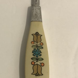 May include: A vintage metal and plastic potato peeler with a white handle featuring a floral design in orange, blue, and green.
