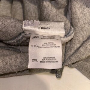 May include: Close-up of gray fabric with multiple tags. The tags include the Disney logo, size labels (2EG, 2TG, 2XL), and fabric composition details: 50% cotton, 50% polyester. The tags also indicate the item was made in Honduras.