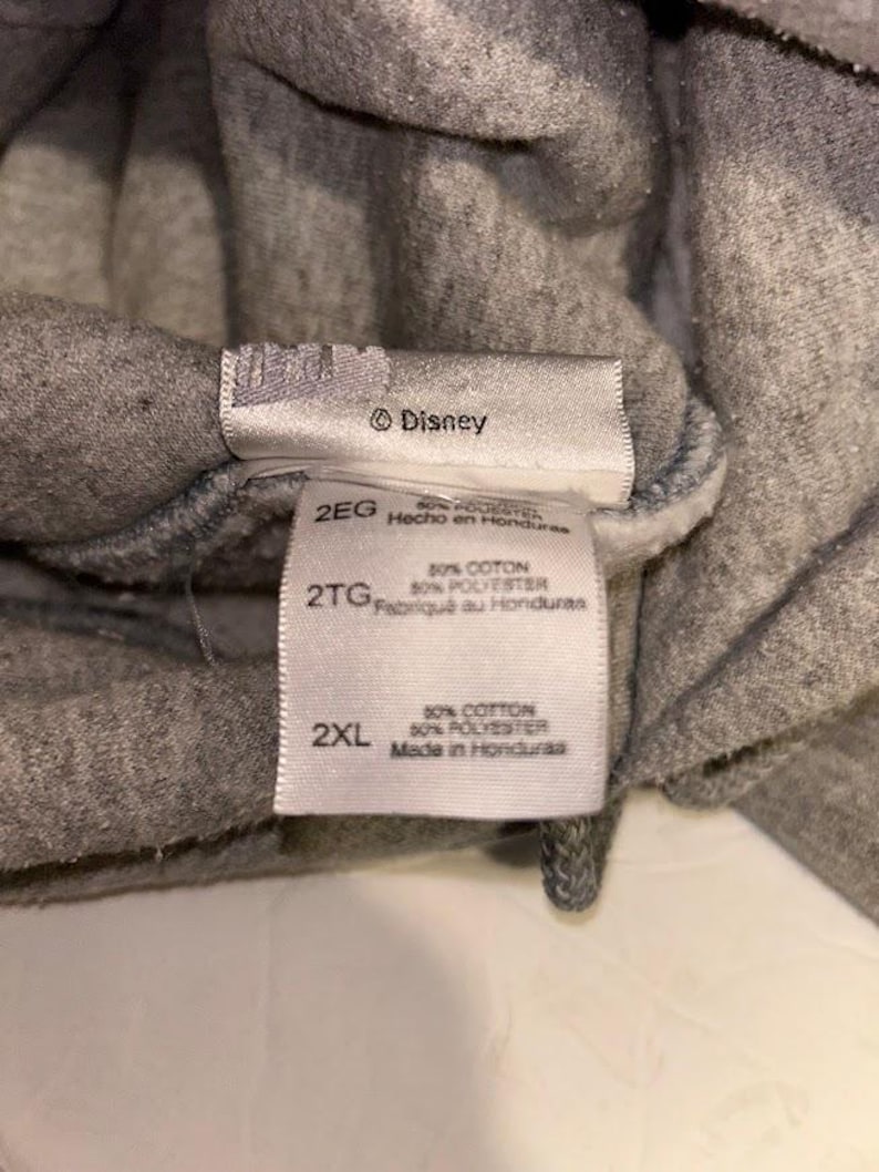 May include: Close-up of gray fabric with multiple tags. The tags include the Disney logo, size labels (2EG, 2TG, 2XL), and fabric composition details: 50% cotton, 50% polyester. The tags also indicate the item was made in Honduras.