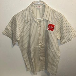 May include: Cream-colored short-sleeve button-up shirt with thin vertical black stripes. The shirt features a Coca-Cola logo patch on the chest pocket. The shirt is hanging on a white hanger.