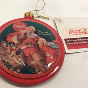 May include: Red Coca-Cola Christmas ornament featuring a vintage illustration of Santa Claus sitting in a chair with a bottle of Coca-Cola and a cooler that says "ICE COLD". The text "Thirst asks nothing more" is printed on the ornament. The ornament comes with a white tag that says "Coca-Cola Christmas Ornament Décoration de Noël".