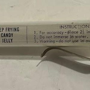 May include: A clear glass deep frying thermometer with a red bulb and white plastic cap. The thermometer has a metal clip and the text "deu-rite DEEP FRYING CANDY JELLY" is printed on the side. Instructions are also printed on the side.