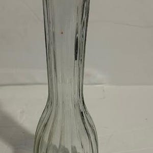 Puede incluir: Un jarrón de vidrio transparente con detalles acanalados verticales. El jarrón tiene una base bulbosa que se estrecha hacia un cuello delgado y un borde acampanado. El jarrón mide aproximadamente 25 cm de alto, adecuado para exhibir flores.