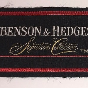 May include: A black fabric patch with red stitching around the edges. The patch features white text that reads "Benson & Hedges Signature Collection" with a gold "TM" symbol.