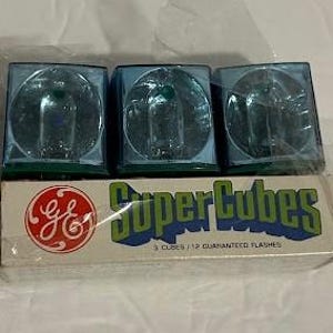 May include: Three blue Super Cubes flash cubes are stacked on top of their original box. The box has the GE logo and the words "Super Cubes" in green and blue. The box reads "3 Cubes / 12 Guaranteed Flashes".