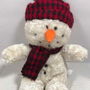 May include: White plush snowman wearing a red and black plaid hat and scarf. The snowman has black eyes, a black mouth, and an orange carrot nose.